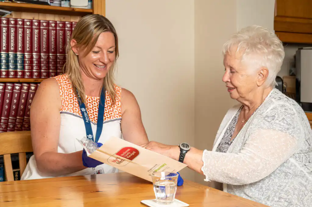 Elderly client being assisted by their professional carer to take her medication. They are looking at a dosset box and there is a full glass of water on the table.