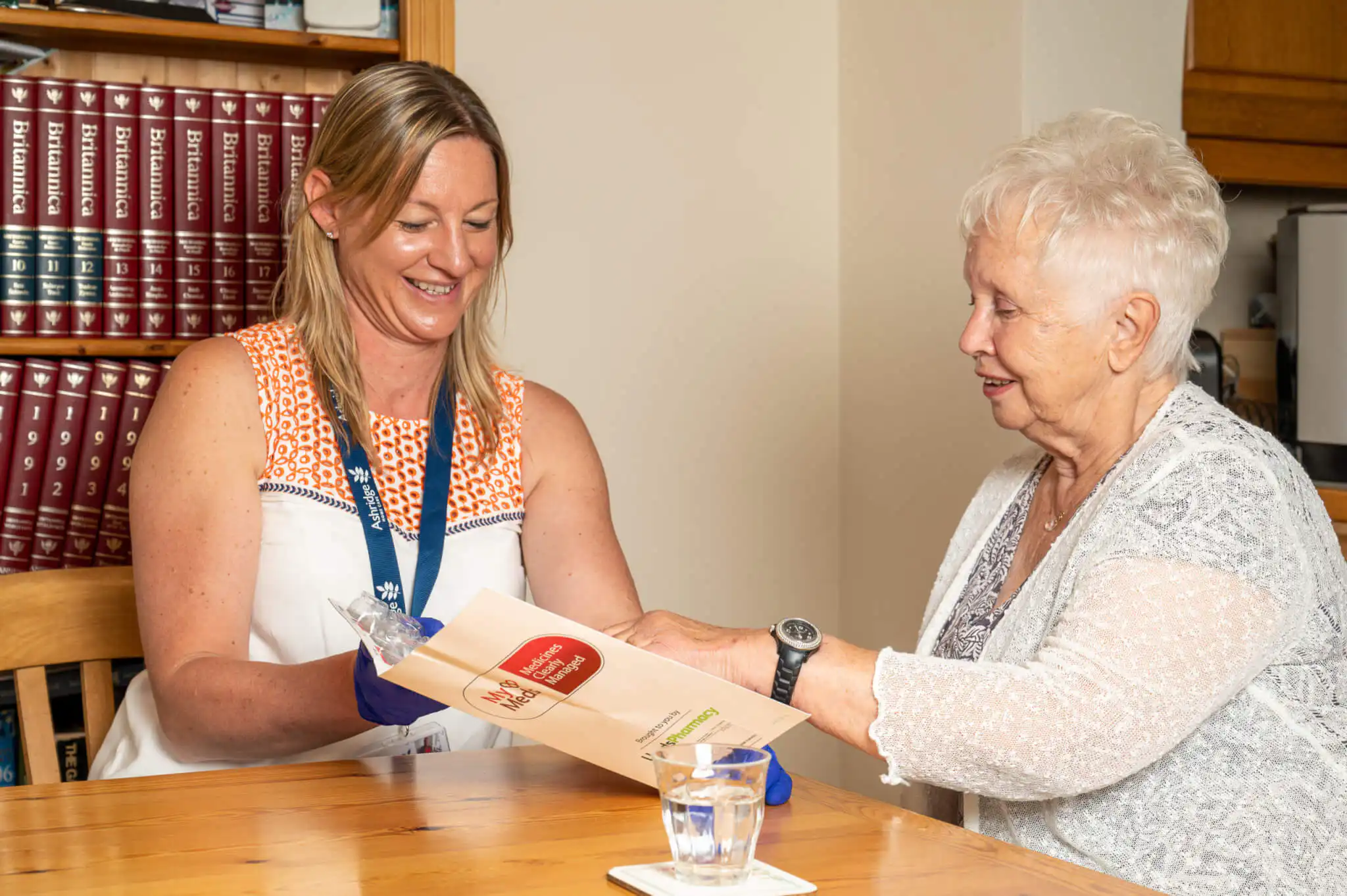 Elderly client being assisted by their professional carer to take her medication. They are looking at a dosset box and there is a full glass of water on the table.