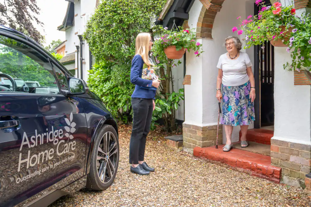 Client services manager Agata greeting a new client at their door ahead of completing their personalised care needs assessment. She is standing in their driveway with an Ashridge Home Care brochure, next to a blue car with Ashridge Home Care branding.