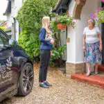 Client services manager Agata greeting a new client at their door ahead of completing their personalised care needs assessment. She is standing in their driveway with an Ashridge Home Care brochure, next to a blue car with Ashridge Home Care branding.