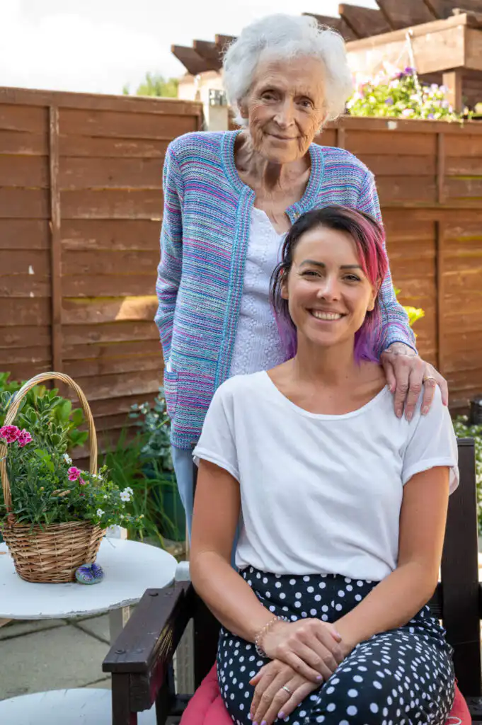 Elderly woman stood behind her granddaughter, resting a hand on her shoulder. Both are in the garden smiling warmly at the camera.
