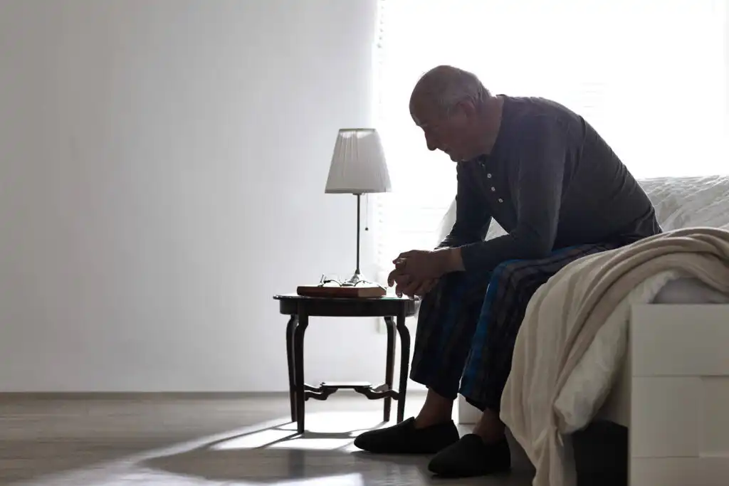 Elderly man sitting on the edge of his bed, he is staring off into the distance looking thoughtful.