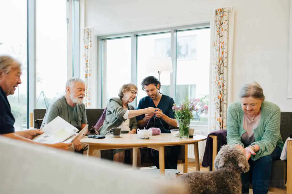 Group of five people sat in a living room enjoying different activities, one person is reading the newspaper, two people are knitting together, one is petting a dog and the other is watching them smiling.