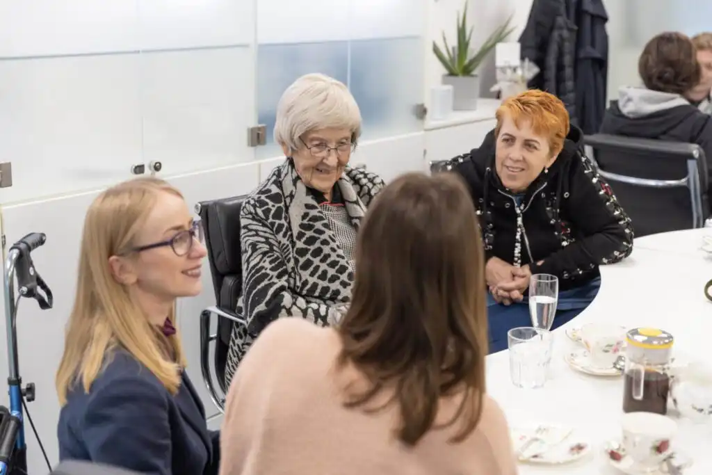 A group of carers, clients and the client services manager sitting around a table enjoying tea and cake during an event at the Ashridge Home Care offices.