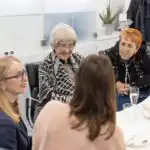 A group of carers, clients and the client services manager sitting around a table enjoying tea and cake during an event at the Ashridge Home Care offices.