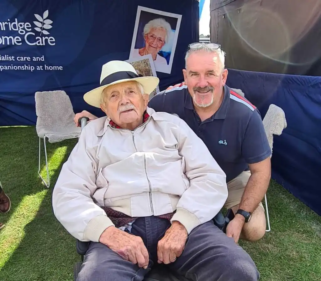 A live-in caregiver assists an elderly man at a community event.