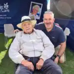 A live-in caregiver assists an elderly man at a community event.