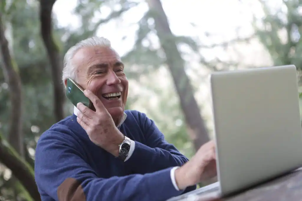 Man sitting at a bench in a park with his laptop whilst laughing on the phone.