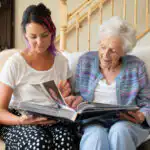 Elderly lady being supported by her carer to look through old photo albums. They are both smiling and look to be enjoying sharing memories.