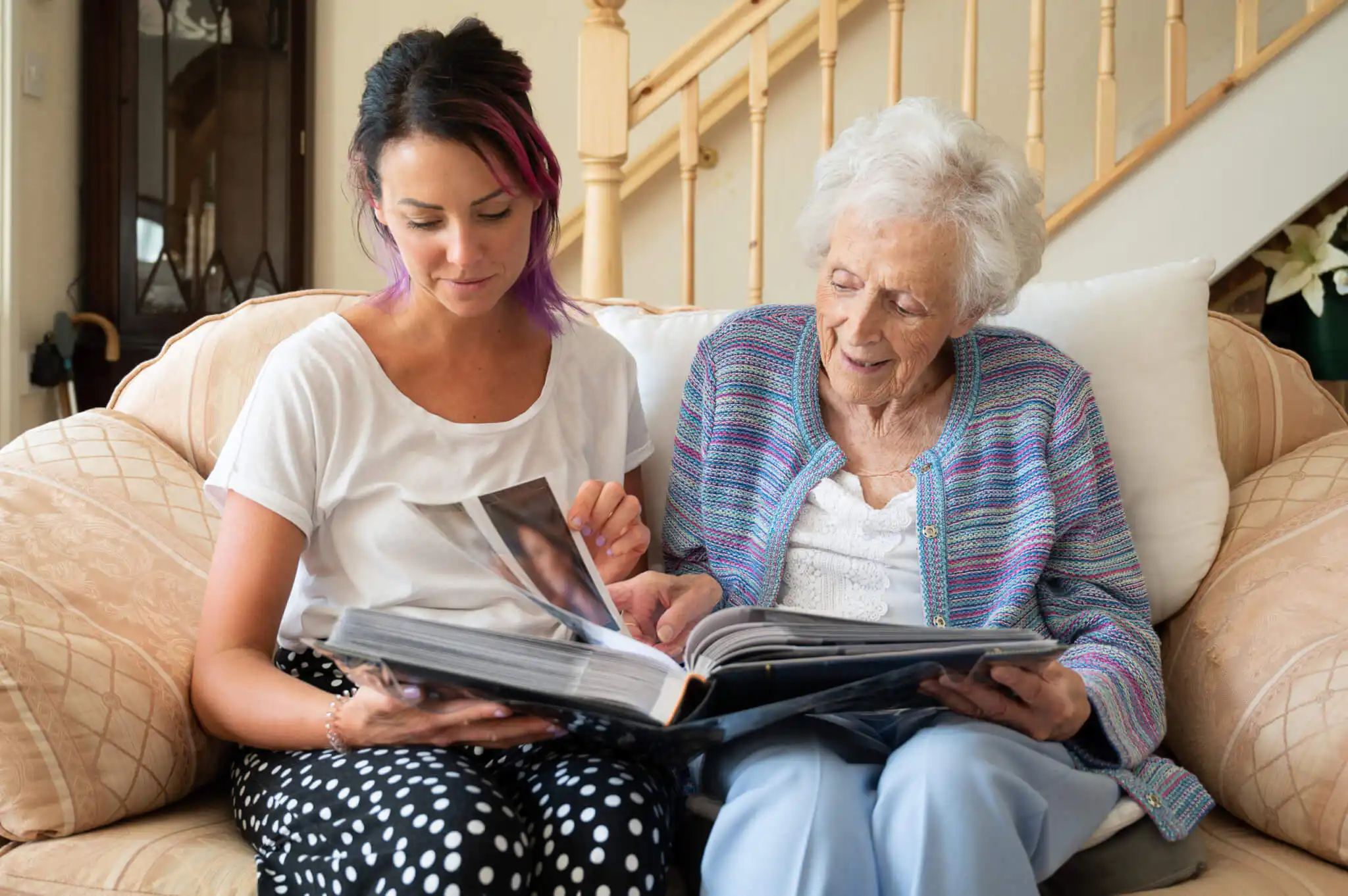 Elderly lady being supported by her carer to look through old photo albums. They are both smiling and look to be enjoying sharing memories.