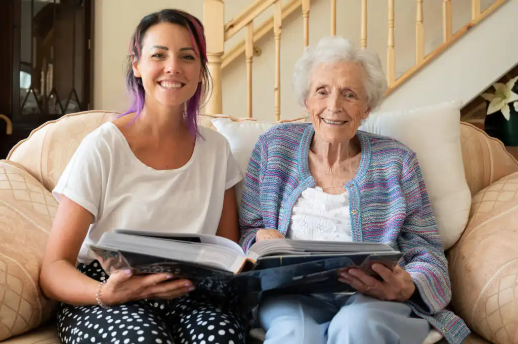 Elderly lady being supported by her carer to look through old photo albums. They are both smiling and look to be enjoying sharing memories.