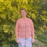 A woman stands outdoors in front of lush green foliage, wearing a pastel pink lace cardigan and light floral pants, smiling warmly at the camera.