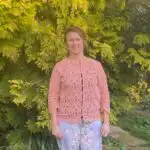 A woman stands outdoors in front of lush green foliage, wearing a pastel pink lace cardigan and light floral pants, smiling warmly at the camera.