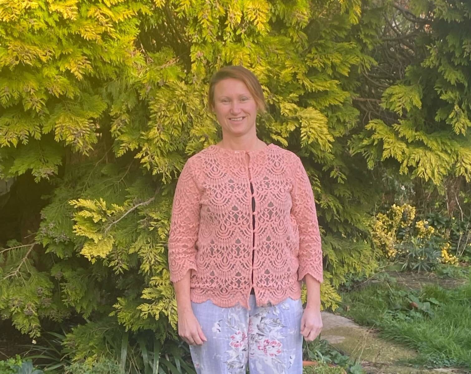 A woman stands outdoors in front of lush green foliage, wearing a pastel pink lace cardigan and light floral pants, smiling warmly at the camera.