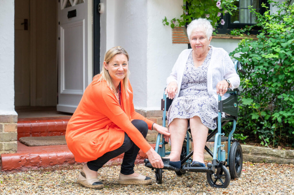 A woman in an orange sweater kneels to adjust a resting elderly woman's wheelchair outside a house. The scene is caring and supportive.