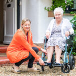 A woman in an orange sweater kneels to adjust a resting elderly woman's wheelchair outside a house. The scene is caring and supportive.