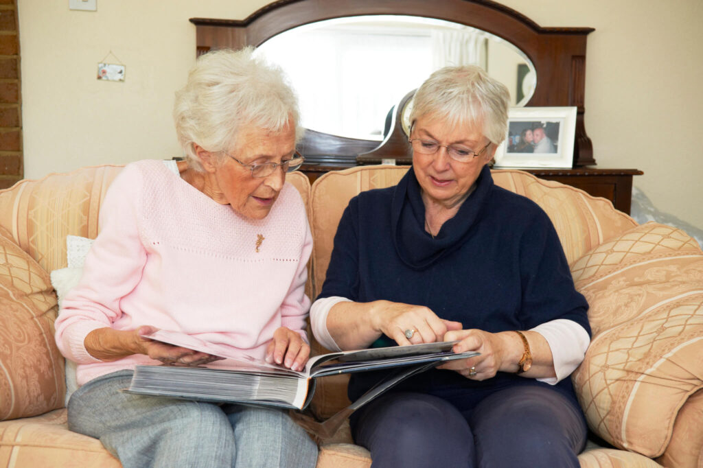 Two elderly women sit on a sofa, sharing a photo album. They wear glasses and cozy sweaters, appearing engaged and nostalgic.