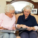 Two elderly women sit on a sofa, sharing a photo album. They wear glasses and cozy sweaters, appearing engaged and nostalgic.