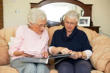 Two elderly women sit on a sofa, sharing a photo album. They wear glasses and cozy sweaters, appearing engaged and nostalgic.