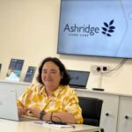 A person in a yellow patterned shirt sits at a desk with a laptop, smiling. Behind, a screen displays "Ashridge Home Care."