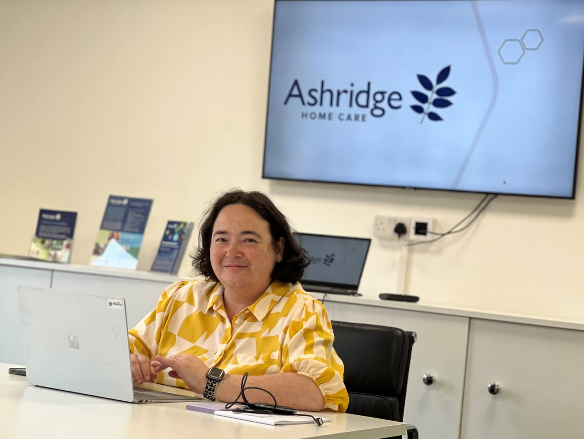 A person in a yellow patterned shirt sits at a desk with a laptop, smiling. Behind, a screen displays "Ashridge Home Care."