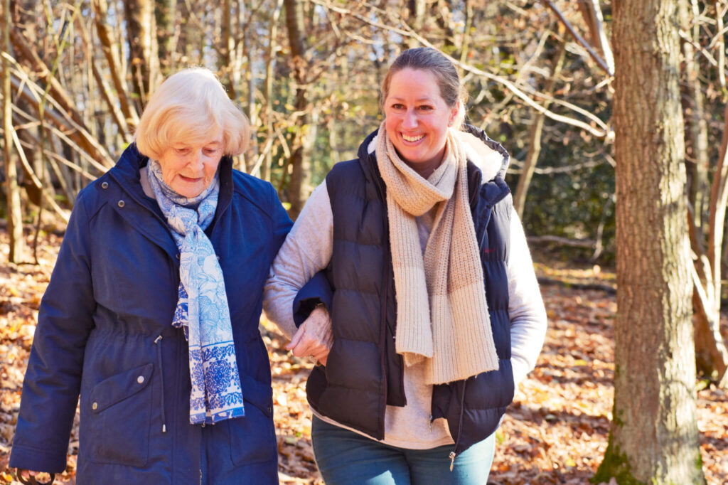 A woman and an elderly woman walk arm in arm through a sunlit, autumnal forest. They are warmly dressed, smiling, and enjoying a peaceful moment.