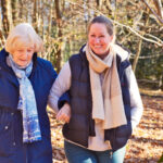 A woman and an elderly woman walk arm in arm through a sunlit, autumnal forest. They are warmly dressed, smiling, and enjoying a peaceful moment.