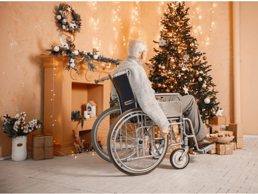 An elderly person in a wheelchair looks at a decorated Christmas tree and a mantel adorned with lights and wreaths.