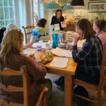 A group of six people sit around a wooden table covered with papers, sharing a meal. One person stands at the far end, holding books.