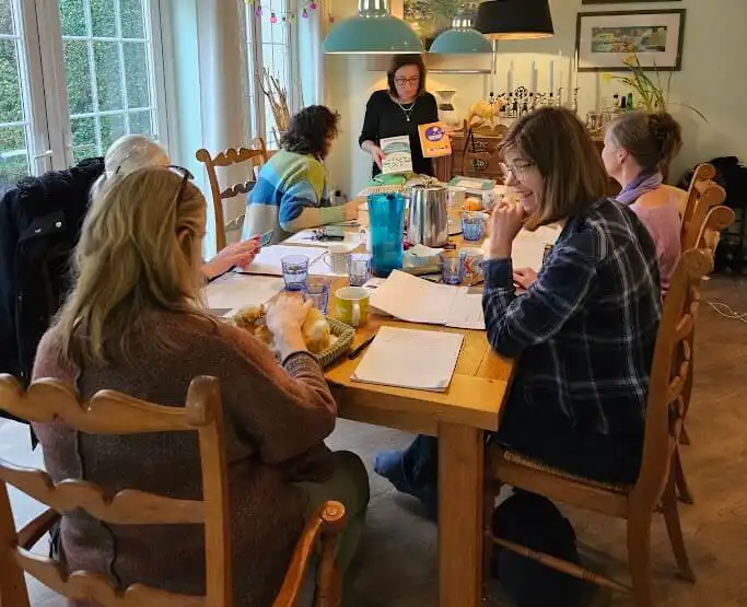 A group of six people sit around a wooden table covered with papers, sharing a meal. One person stands at the far end, holding books.