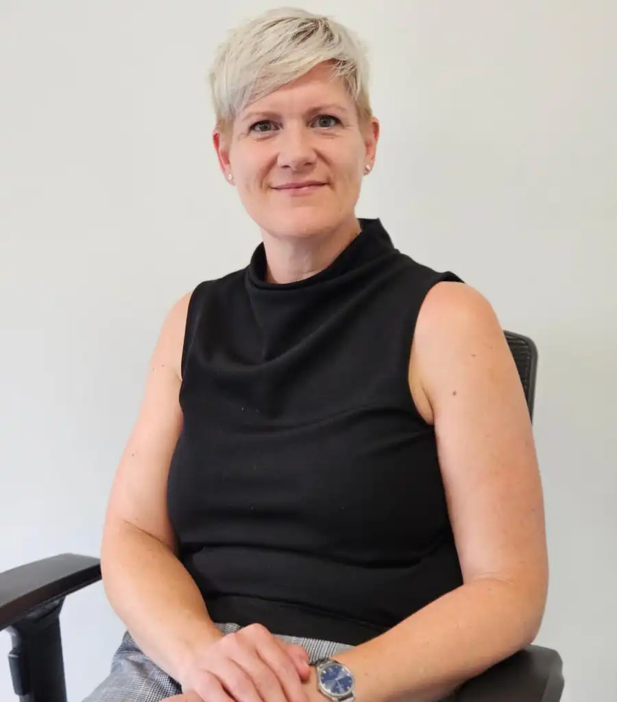 A person with short light hair sits confidently in a black sleeveless top, hands resting in their lap, against a plain light grey background.