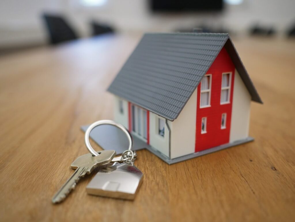 A small model house with a red and white facade sits on a wooden table next to a set of silver keys, symbolising homeownership or real estate.