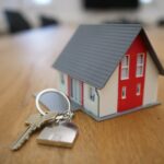 A small model house with a red and white facade sits on a wooden table next to a set of silver keys, symbolising homeownership or real estate.