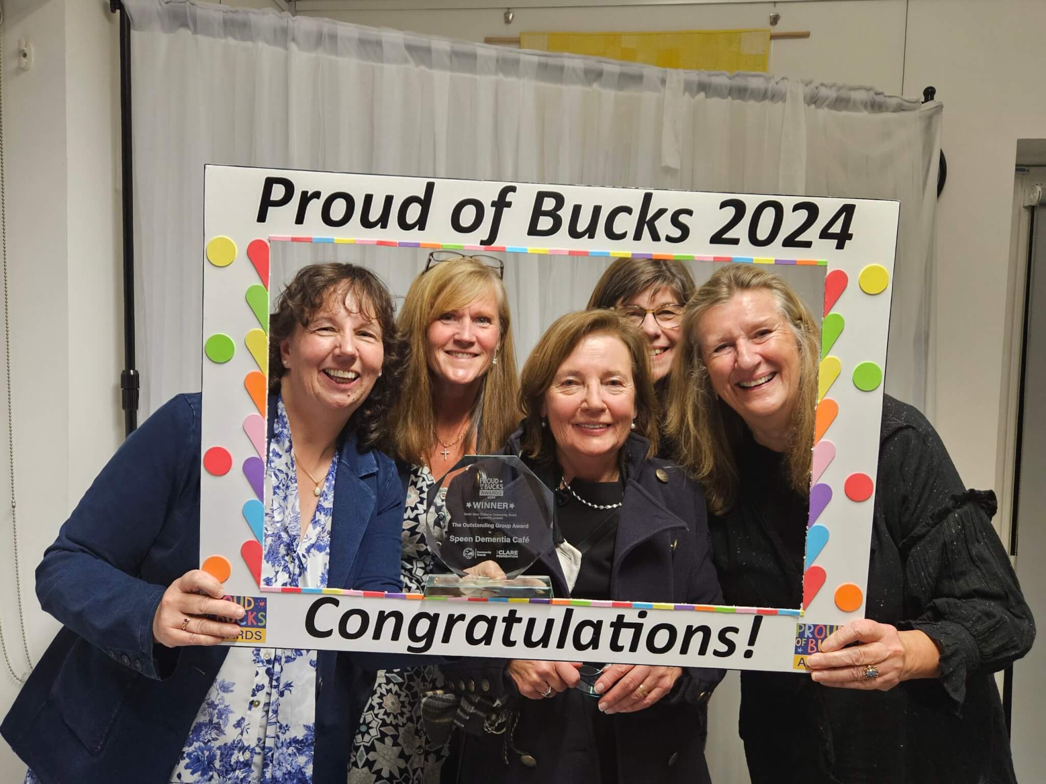 Five smiling people hold a colourful "Proud of Bucks 2024" frame. The person in front holds an award.