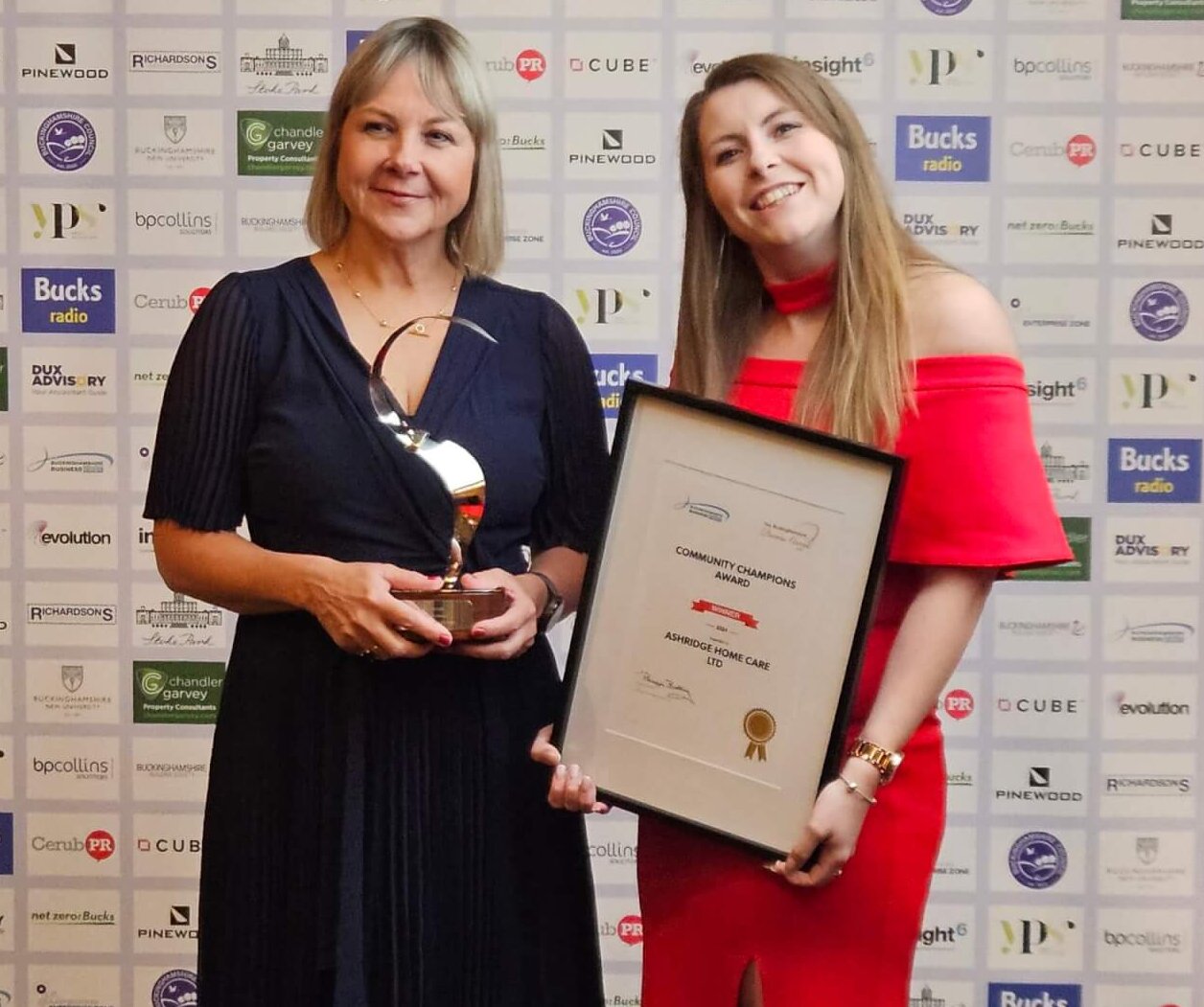 Two women smiling proudly, one holding a trophy and the other holding a framed award. They stand against a backdrop with multiple sponsor logos.