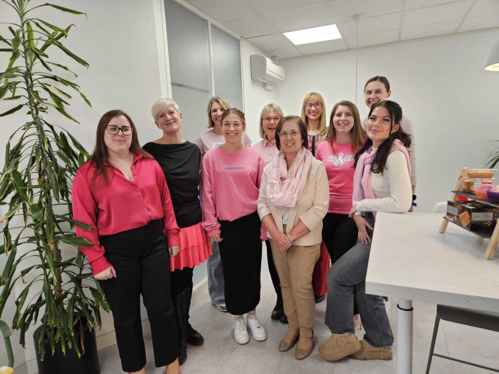 A group of ten smiling people stands in an office wearing pink and white attire. A tall plant and office furniture create a friendly, casual atmosphere.