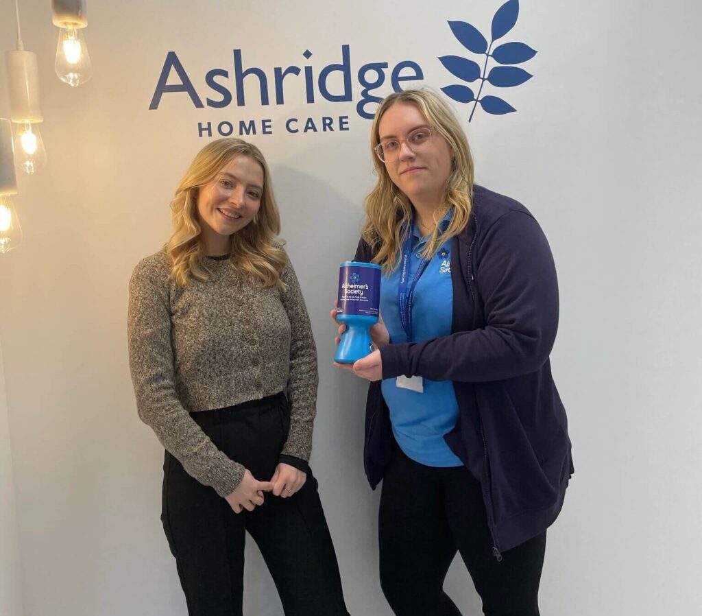 Two women stand smiling in front of an Ashridge Home Care logo. One holds a blue charity donation box. The setting is bright with hanging lights.