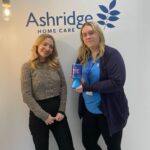 Two women stand smiling in front of an Ashridge Home Care logo. One holds a blue charity donation box. The setting is bright with hanging lights.