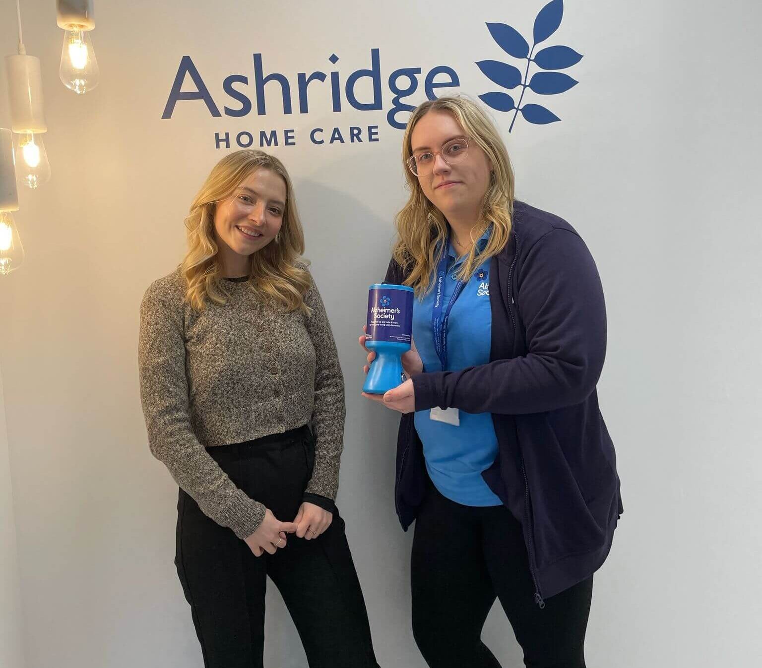 Two women stand smiling in front of an Ashridge Home Care logo. One holds a blue charity donation box. The setting is bright with hanging lights.