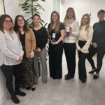 Seven women standing in an office, smiling, wearing smart casual attire. Two hold award plaques. A large plant is visible in the background.