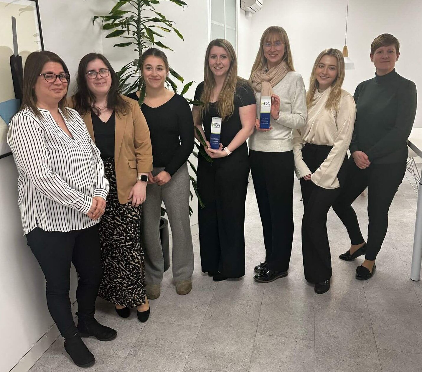 Seven women standing in an office, smiling, wearing smart casual attire. Two hold award plaques. A large plant is visible in the background.