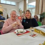 Two women sit at a table in a cozy room, smiling warmly. One holds a napkin, while the other wears a dark sweater.