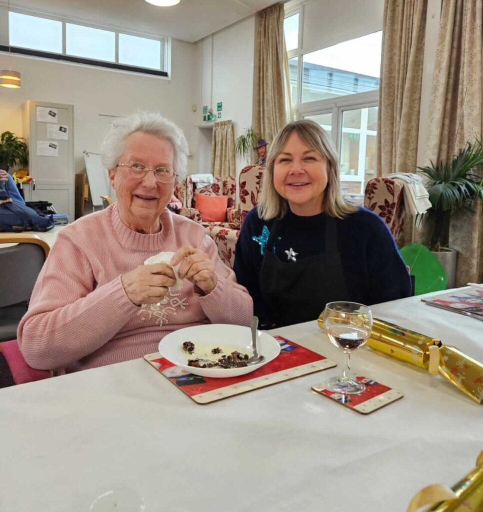 Two women sit at a table in a cozy room, smiling warmly. One holds a napkin, while the other wears a dark sweater.