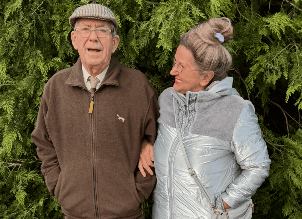Elderly couple standing together, smiling warmly. The man wears a flat cap and brown jacket, while the woman in a silver coat glances at him.