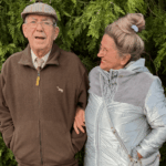 Elderly couple standing together, smiling warmly. The man wears a flat cap and brown jacket, while the woman in a silver coat glances at him.