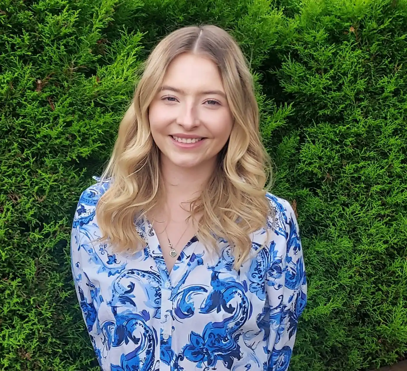 A person with long blonde hair smiles warmly while standing in front of dense green foliage. They wear a blue and white patterned shirt.