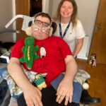 A smiling woman stands beside a man in a wheelchair wearing a red cartoon shirt and breathing apparatus. Plush toys rest on his lap.