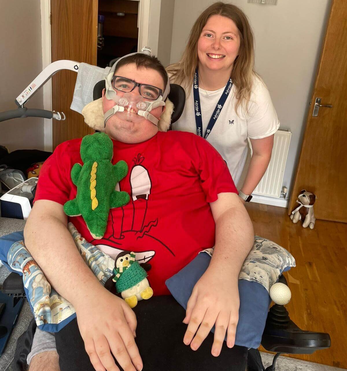 A smiling woman stands beside a man in a wheelchair wearing a red cartoon shirt and breathing apparatus. Plush toys rest on his lap.