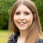 A woman with long brown hair smiles warmly at the camera. She is outdoors, standing in front of a blurred green background, wearing a polka-dot top.