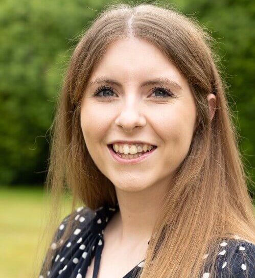 A woman with long brown hair smiles warmly at the camera. She is outdoors, standing in front of a blurred green background, wearing a polka-dot top.